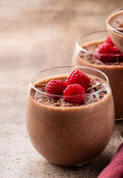 Homemade chocolate mousse decorated with raspberries and dark chocolate sprinkles in portion glasses. Brown table. Selective focus, copy space.