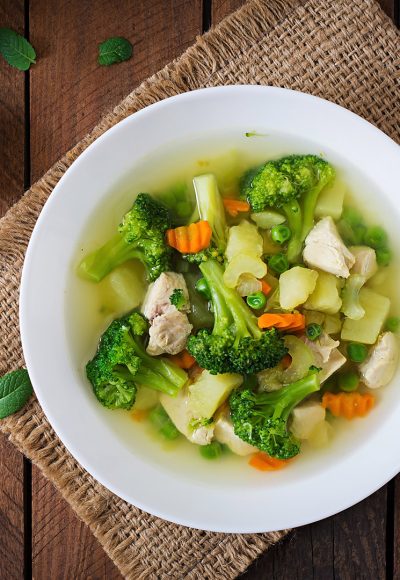 Chicken soup with broccoli, green peas, carrots and celery in a white bowl on a wooden background in rustic style. Top view