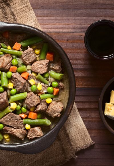 Beef stew or soup with colorful summer vegetables (pea, carrot, sweet corn, green bean, onion) in rustic bowl with red wine and bread slices on the side, photographed overhead with natural light