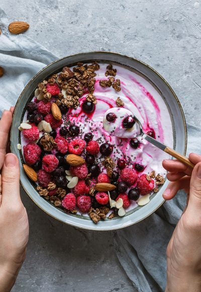 Hands holding a bowl of natural yogurt with oat granola, frozen raspberries, blueberries, nuts and seeds in bowl on light background for healthy breakfast. Healthy detox smoothy bowl, dieting