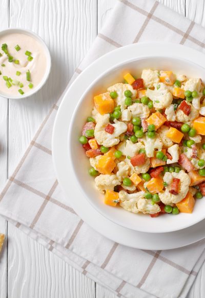 cauliflower salad with crispy fried bacon, green peas and cheddar cheese cubes in a white bowl on a wooden table with golden cutlery, view from above, flatlay