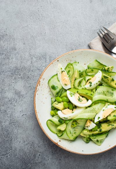 Healthy green avocado salad bowl with boiled eggs, sliced cucumbers, edamame beans, olive oil and herbs on ceramic plate top view, grey stone rustic table background. Copy space