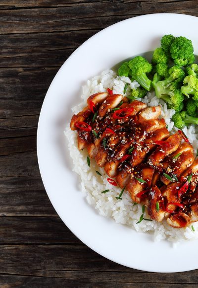 white rice topped with teriyaki chicken breast sprinkled with chives, sesame seeds, finely chopped chili peppers served with steamed broccoli on white plate, view from above