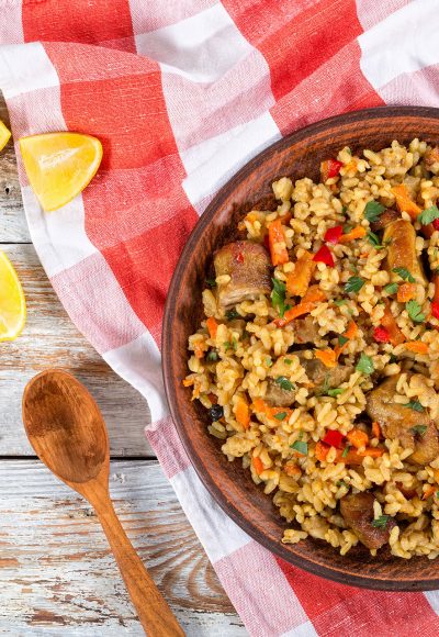 rice with meat, pepper, vegetables and spices on dish, lemon slice, garlic, chili pepper and wooden spoon on white peeling paint planks, view from above