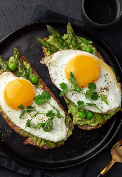 toasts with avocado, asparagus and fried egg on black plate, top view, dark background, copy space