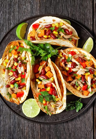 tacos of flour tortilla shells filling with grilled chicken meat, corn, roasted sweet potatoes cubes, red pepper and parsley served on a black plate, flat lay, free space