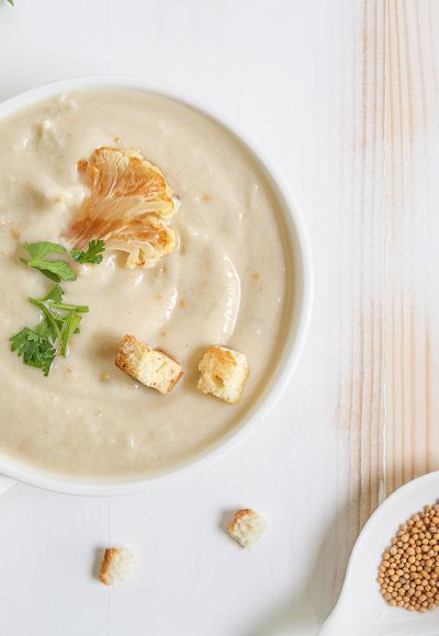 Homemade white cauliflower cream soup with fresh shopped green herbs and croutons in white ceramic bowl on wooden white and beige table. Horizontal orientation. Flat lay. Copy space.