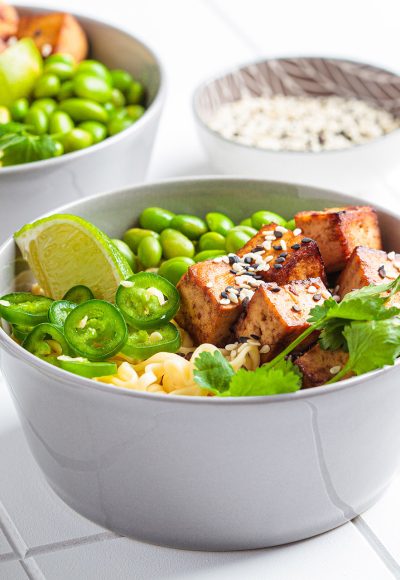 Vegan noodles ramen soup with marinated tofu, edamame beans and hot peppers in a gray bowl on a white tile background.